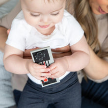 Load image into Gallery viewer, A baby holding a Bumkins Silicone Teether: NES™ Controller sits on a woman's lap.