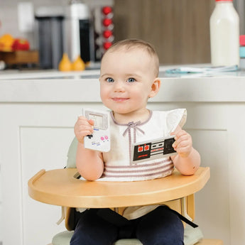 A baby in a wood high chair holds Bumkins Silicone Teether: Game Boy™ and another teether in a kitchen.