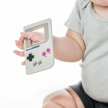 Close-up of a baby in a gray outfit holding a Bumkins Silicone Teether: Game Boy™ on a white background.