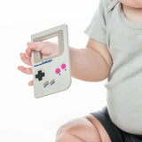 Close-up of a baby in a gray outfit holding a Bumkins Silicone Teether: Game Boy™ on a white background.