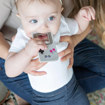 A seated adult holds a child chewing a Bumkins Silicone Teether: Game Boy™.