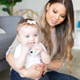 A woman holds a baby happily chewing on a Bumkins Silicone Teether: Game Boy™ while sitting in a cozy living room.