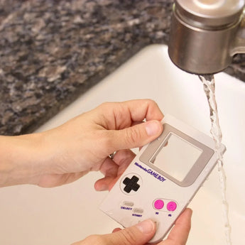 Hands washing a Bumkins Silicone Teether: Game Boy™ under a running kitchen sink faucet.
