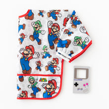 Load image into Gallery viewer, Bumkins Sleeved Bib: Super Mario™ Classic lays folded on a white background next to a gray GameBoy-shaped silicone teether.