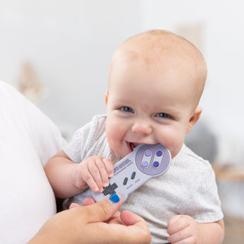A baby is playfully biting a Bumkins Silicone Teether: Nintendo SNES™ Controller while held by someone with blue-painted nails.