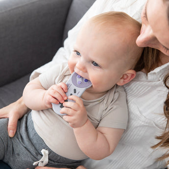 A baby chews a Bumkins Silicone Teether: Nintendo SNES™ Controller while sitting on an adult's lap on a gray couch.