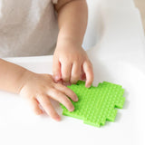 Close-up of a child's hands grasping a face-down Bumkins Silicone Teether: Zelda™ Link on a high chair tray.