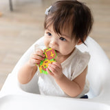 A toddler in a high chair, wearing a white shirt, chews a Bumkins Silicone Teether: Zelda™ Link.