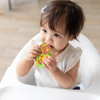 A toddler in a high chair, wearing a white shirt, chews a Bumkins Silicone Teether: Zelda™ Link.