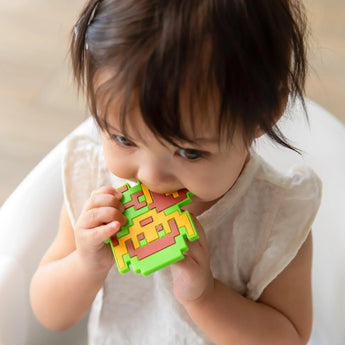 A toddler in white shirt in a high chair chews a Bumkins Silicone Teether: Zelda™ Link.