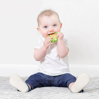 A baby on a carpet in white and blue clothes chews a Bumkins Silicone Teether: Zelda™ Link.
