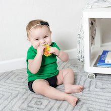 Load image into Gallery viewer, A baby sits on a gray carpet, smiling and chewing a Bumkins Silicone Teether: Zelda™ Link near a white shelf.