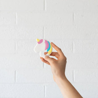 A hand holds a Bumkins Silicone Teether: Unicorn against a white brick wall.