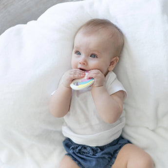 A baby in a white shirt and blue shorts sits on a white blanket, chewing a Bumkins Silicone Teether: Unicorn.