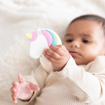A baby in a beige outfit holds a Bumkins Silicone Teether: Unicorn on a soft, light-colored blanket.