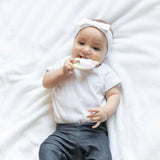 A baby in a headband and white onesie on a soft blanket holds a Bumkins Silicone Teether: Unicorn.