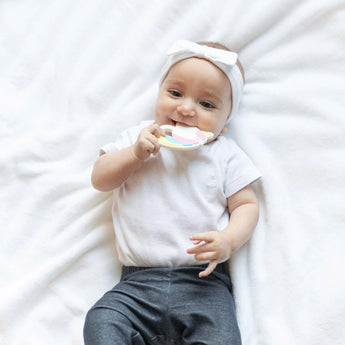 A baby in a headband and white onesie on a soft blanket holds a Bumkins Silicone Teether: Unicorn.