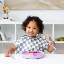 Load image into Gallery viewer, A curly-haired toddler smiles, wearing a Bumkins Junior Bib: Charcoal Check while eating from a purple plate with utensils at a white table.