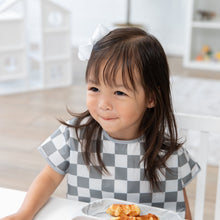 Load image into Gallery viewer, A young girl in a Bumkins Junior Bib: Charcoal Check sits at a table with waffles, smiling softly.
