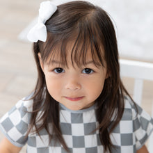 Load image into Gallery viewer, A young girl with brown hair and a bow smiles in a Bumkins Junior Bib: Charcoal Check, made from waterproof fabric.