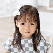Load image into Gallery viewer, A young girl sits and smiles in a bright room, wearing a Bumkins Junior Bib: Charcoal Check with waterproof, easy-wipe fabric.