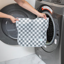 Load image into Gallery viewer, A person holds Bumkins Junior Bib: Charcoal Check in front of an open front-loading washing machine.