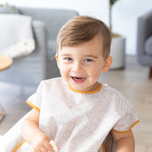 Load image into Gallery viewer, A happy toddler sits in a highchair wearing a Bumkins Junior Bib: Wander. A blurry living room with a couch and plant is in the background.