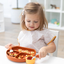 Load image into Gallery viewer, A toddler wears a Bumkins Junior Bib: Wander, while eating from a divided plate with a spoon at a white tablee.