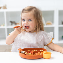 Load image into Gallery viewer, A toddler enjoys a meal using a spoon while wearing a Bumkins Junior Bib: Wander, eating from a colorful tray at a white table.