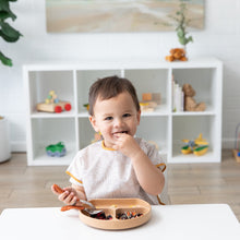 Load image into Gallery viewer, A toddler at a white table wears a Bumkins Junior Bib: Wander, smiling while eating from a divided plate, with toys on a white shelf behind.