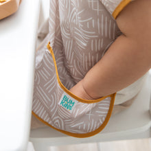 Load image into Gallery viewer, Close-up of a child at a white table, with their hand nestled in the catch-all pocket of a Bumkins Junior Bib: Wander.