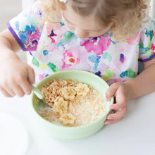 Load image into Gallery viewer, A child wearing a Bumkins Junior Bib: Watercolor enjoys cereal with milk and bananas from a green bowl with a spoon.