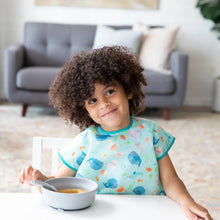 Load image into Gallery viewer, A curly-haired child smiles, wearing a Bumkins Junior Bib: Ocean Life, with a gray bowl and spoon on a white table.