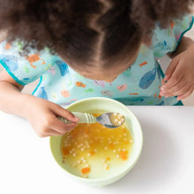 Load image into Gallery viewer, A curly-haired child wearing a Bumkins Junior Bib: Ocean Life sits at a white table, eating soup from a light green bowl.