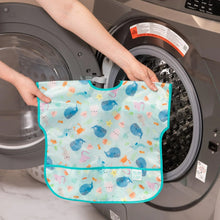 Load image into Gallery viewer, A person holds Bumkins Junior Bib: Ocean Life in front of an open front-loading washing machine.