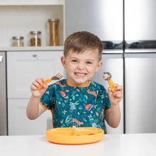 Load image into Gallery viewer, A child in a colorful Bumkins Junior Bib: Jungle smiles, holding a fork and spoon at a table beside an orange plate.