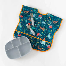 Load image into Gallery viewer, Bumkins Junior Bib: Jungle lays flat on a white background with gray utensils on top and a gray divided plate nearby.
