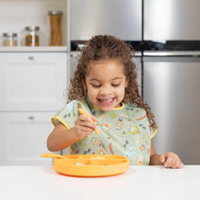 Load image into Gallery viewer, A child smiles while using a fork to eat from an orange plate, wearing a Bumkins Junior Bib: Camp Gear at a white table.