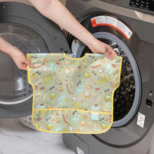 Load image into Gallery viewer, A person holds Bumkins Junior Bib: Camp Gear in front of an open front-loading washing machine.