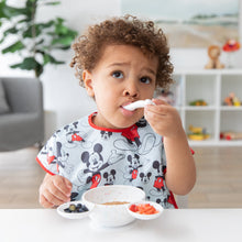 Load image into Gallery viewer, A young child in a Bumkins Junior Bib: Mickey Mouse Classic eats oatmeal, blueberries, and strawberries from a white bowl at a white table.