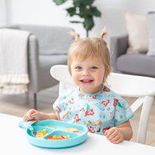 Load image into Gallery viewer, A smiling toddler in pigtails wears a Bumkins Junior Bib: Ariel and eats happily from a divided plate with utensils at a table.