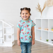 Load image into Gallery viewer, A girl with pigtails wearing a Bumkins Junior Bib: Ariel smiles, standing in front of a dollhouse and toy-filled shelves.