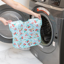 Load image into Gallery viewer, A person holds a Bumkins Junior Bib: Ariel, featuring cartoon designs and waterproof fabric, in front of a front-loading washing machine.
