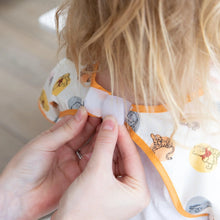 Load image into Gallery viewer, Close-up of hands fastening the hook-and-loop closure of a Bumkins Junior Bib: Winnie and Friends around a child's neck.