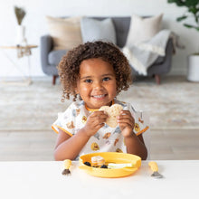 Load image into Gallery viewer, A child beams, holding a sandwich by a yellow plate and utensils, wearing a Bumkins Junior Bib: Winnie and Friends in a cozy living room.