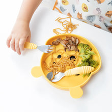 Load image into Gallery viewer, Close-up of a child wearing Bumkins Junior Bib: Winnie and Friends, holding a fork near a bear-shaped plate of food on a white table.