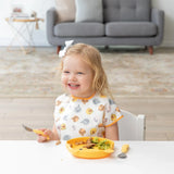 A girl in a Bumkins Junior Bib: Winnie and Friends smiles while eating with a fork from a yellow plate at a white table.