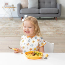 Load image into Gallery viewer, A girl in a Bumkins Junior Bib: Winnie and Friends smiles while eating with a fork from a yellow plate at a white table.