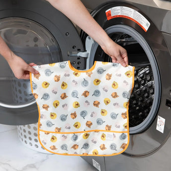 A person holds Bumkins Junior Bib: Winnie and Friends in front of an open front-loading washing machine.