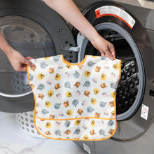 Load image into Gallery viewer, A person holds Bumkins Junior Bib: Winnie and Friends in front of an open front-loading washing machine.